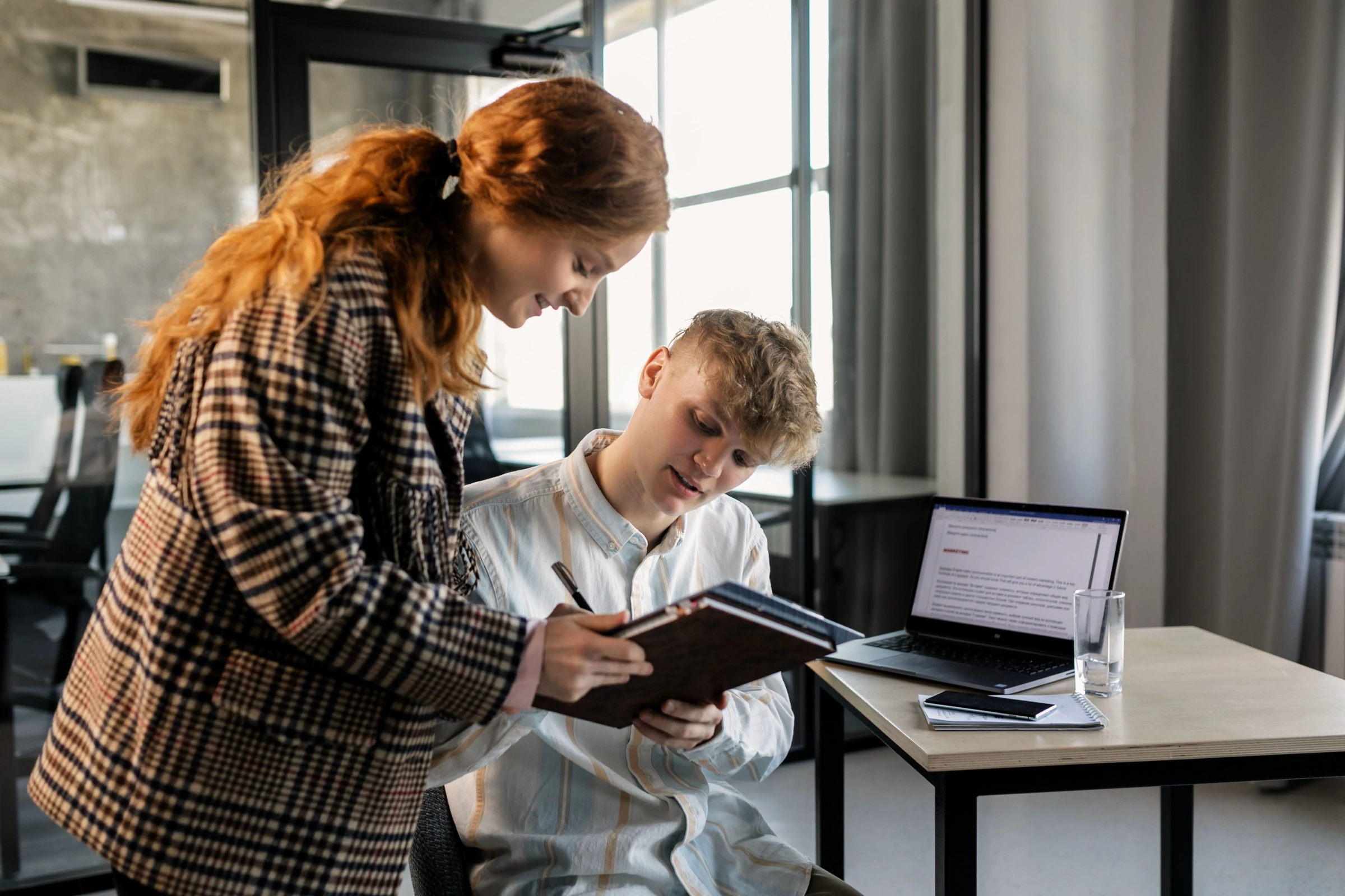 Two colleagues reviewing a notebook together beside a laptop.